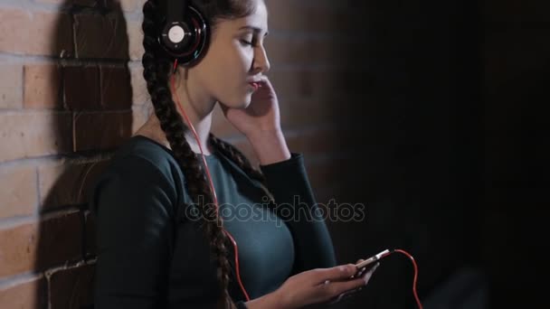 Mujer en auriculares escuchar música en la pared de ladrillo en habitación oscura — Vídeos de Stock