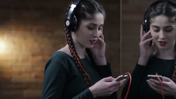 Женщина в наушниках слушает музыку у зеркала в темной комнате — стоковое видео