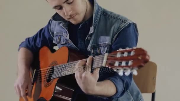 Guitarrista tocando en la guitarra acústica sentado en el fondo de la pared gris — Vídeos de Stock