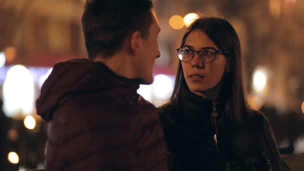 Jong koppel zittend op een bankje samen in een nacht straat — Stockvideo