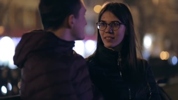 Jong koppel zittend op een bankje samen in een nacht straat — Stockvideo