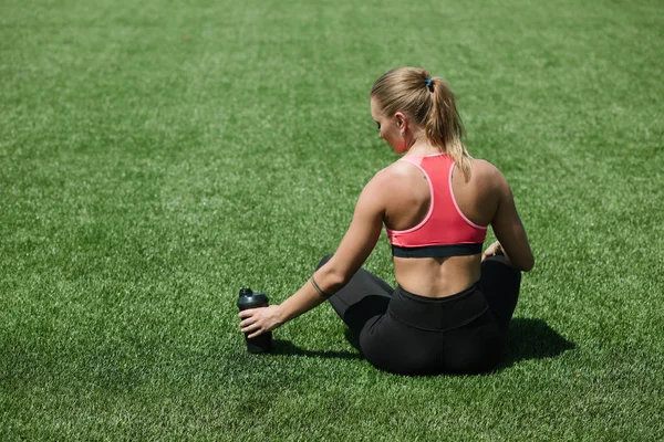 Genç tanınmaz halde sportif kadın atlet spor giyim oturuşta tutmak su veya protein kokteyl sallamak Stadyumu alan yeşil çimenlerin üzerinde egzersiz kavramı — Stok fotoğraf