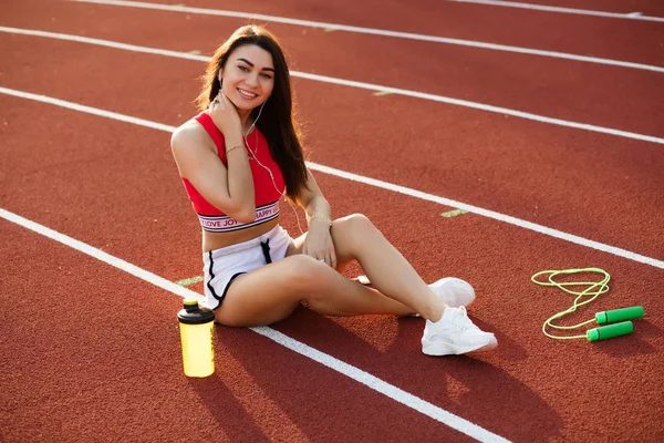 Sexy fitness femminile.Young seducente donna sportiva seduta e sorridere sulla pista dello stadio con scuotimento, concetto di stile di vita sano . — Foto Stock