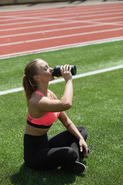 Young sporty woman athlete in sportswear sitting, drinking water or protein cocktail from shake on stadium field green grass, workout concept — Stock Photo, Image