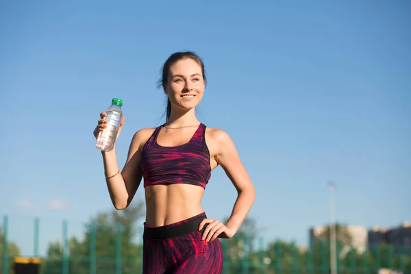 El retrato de la mujer feliz joven guarda la botella con el agua. Deportista sonriente en ropa deportiva sosteniendo bebida en el estadio al aire libre en el fondo del cielo . — Foto de Stock