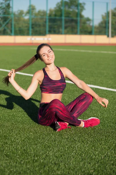 Giovane sportiva sorridente in abbigliamento sportivo seduta sullo stadio erba verde all'aperto. Un concetto di stile di vita sano. Attività sportive estive . — Foto Stock