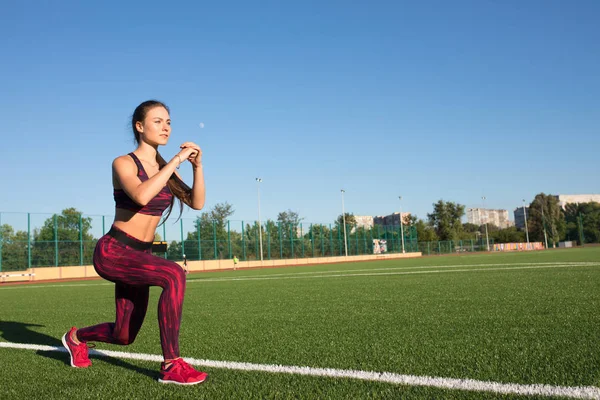 Young happy sportswoman in sportswear making lunges exercise on stadium field area outdoors. Healthy lifestyle concept, sport activity. — Stock Photo, Image