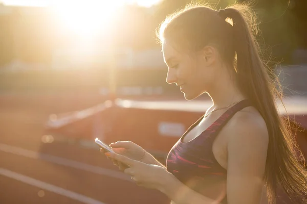 Sportswoman using smart phone outdoors at sunset. Female athlete in sportswear on stadium outdoors. Modern technology concept, sport activity. — Stock Photo, Image