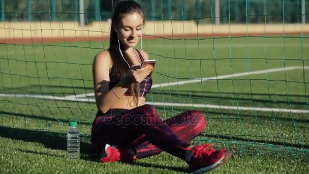 Giovane sportiva in abbigliamento sportivo ascolta musica con auricolari e smartphone seduta sull'erba dello stadio all'aperto. Concetto di stile di vita sano, tecnologie moderne . — Video Stock