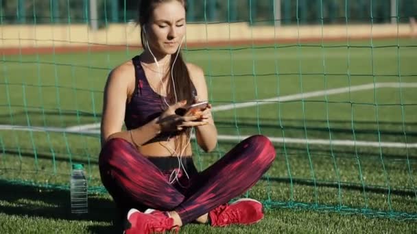 Jovem desportista em sportswear ouvir música com fones de ouvido e telefone inteligente sentado na grama estádio ao ar livre. Conceito de estilo de vida saudável, tecnologias modernas . — Vídeo de Stock