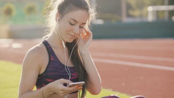 Joven deportista en ropa deportiva escuchar música con auriculares y teléfono inteligente sentado en el estadio al aire libre. Concepto de estilo de vida saludable, tecnologías modernas . — Vídeo de stock