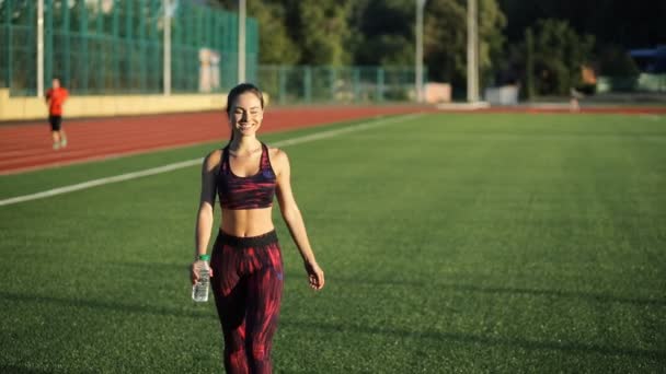 Unga idrottskvinna dricksvatten från flaskan på stadion utomhus. Leende kvinna i sportkläder gå till kamera och innehav dryck. — Stockvideo