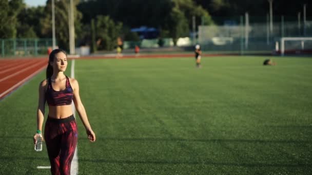 Joven deportista bebiendo agua de la botella en el estadio al aire libre. Sonriente hembra en ropa deportiva ir a la cámara y celebración de la bebida . — Vídeos de Stock