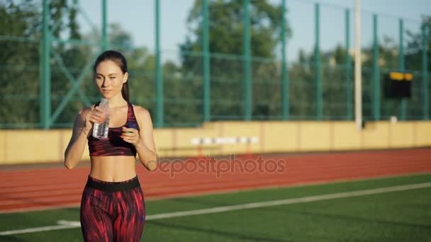 Młody sprawny wody pitnej z butelki na stadion na zewnątrz. Uśmiecha się kobieta w sportowej przejdź do aparatu i napój gospodarstwa. — Wideo stockowe