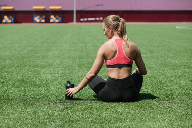 Genç tanınmaz halde sportif kadın atlet spor giyim oturuşta tutmak su veya protein kokteyl sallamak Stadyumu alan yeşil çimenlerin üzerinde egzersiz kavramı