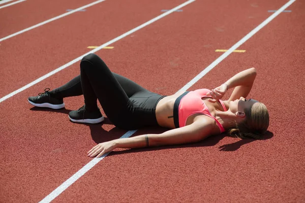 Young tired sporty woman athlete in sportswear lying and relaxing on stadium track after hard workout — Stock Photo, Image