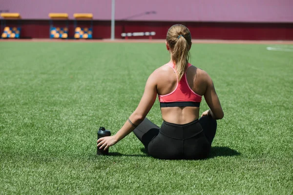 Genç tanınmaz halde sportif kadın atlet spor giyim oturuşta tutmak su veya protein kokteyl sallamak Stadyumu alan yeşil çimenlerin üzerinde egzersiz kavramı — Stok fotoğraf