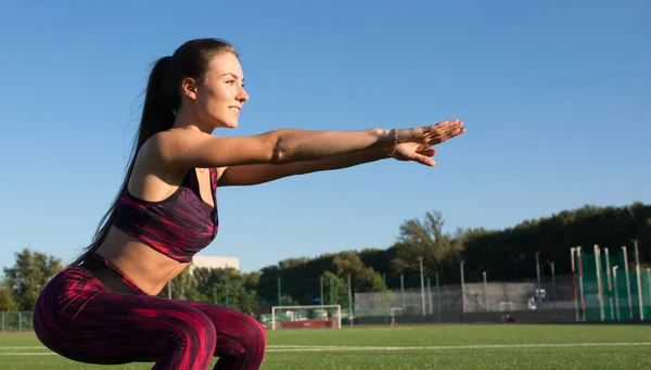 Joven deportista feliz en ropa deportiva haciendo ejercicio en cuclillas en la zona de campo del estadio al aire libre. Concepto de estilo de vida saludable, actividad deportiva . — Foto de Stock