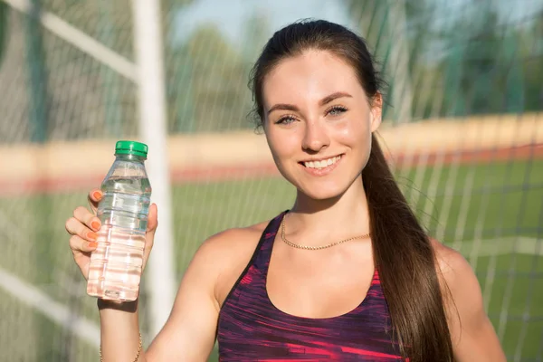 Nahaufnahme Porträt einer jungen glücklichen Frau halten Flasche mit Wasser. Lächelnde Sportlerin in Sportkleidung mit Getränken im Stadion. Konzept eines gesunden Lebensstils. — Stockfoto