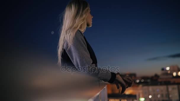 Mujer de pie en la terraza en la ciudad por la noche y mira al paisaje urbano — Vídeo de stock