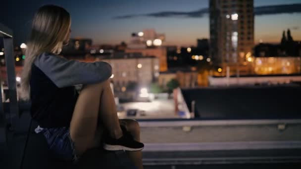 Jeune femme assise sur le toit de la ville la nuit et regarde vers le paysage urbain — Video