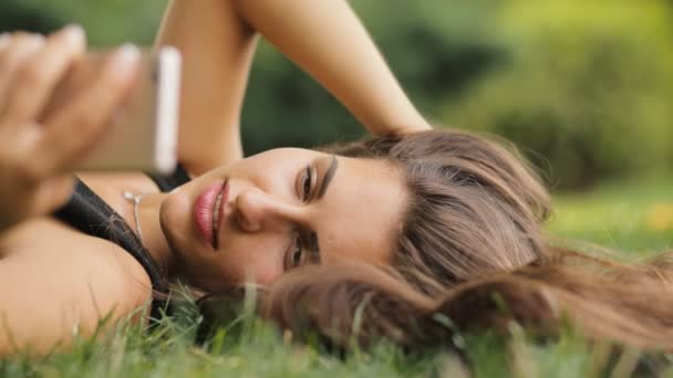 Attraktive Frau im Sommer im Gras liegend, entspannt sie sich und blickt auf den Smartphone-Bildschirm. sie schreibt SMS oder surft im Internet. — Stockvideo