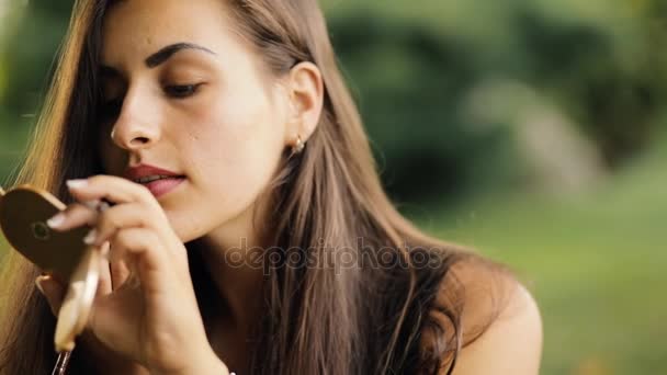 Attractive woman paints lips with pencil sitting on grass in park at summertime, makeup outdoor. — Stock Video