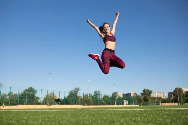 Giovane sportiva felice in abbigliamento sportivo saltare e volare su erba verde stadio all'aperto. Le piace l'estate. Stile di vita sano concetto, attività sportiva . — Foto Stock