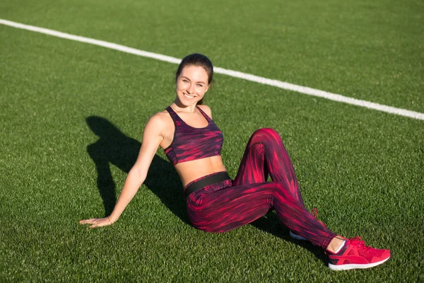 Jonge gelukkig Sportvrouw in sportkleding zittend op stadion groen gras buitenshuis. Gezonde levensstijl concept. Zomer sport activiteit. — Stockfoto