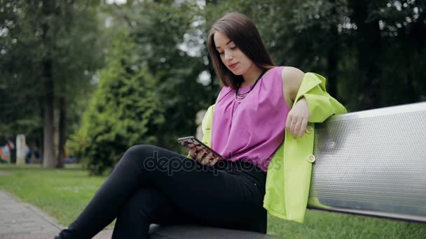 Интернет-зависимость. Женщина сидит на скамейке и использует смартфон в пасмурный летний или осенний день . — стоковое видео