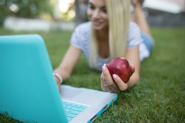 Mulher trabalhando com seu laptop ao ar livre deitado na grama no parque, ela manter uma grande maçã vermelha — Fotografia de Stock
