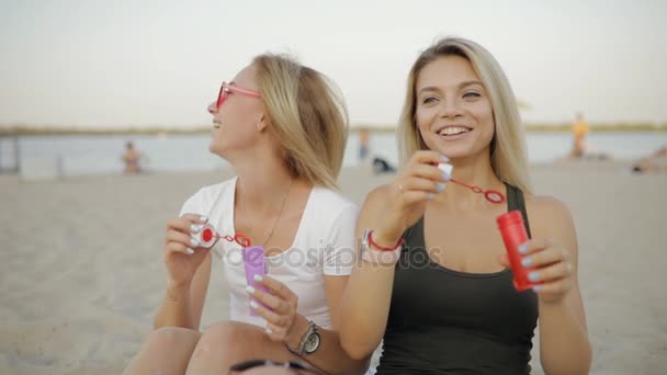 Meninas melhores amigos golpes sabão bolhas brinquedo sentado em uma praia descansando juntos em férias de verão em câmera lenta — Vídeo de Stock