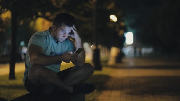 Hombre sentado en un banco en la ciudad nocturna y usando un teléfono inteligente — Vídeos de Stock