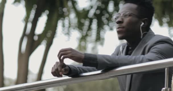 屋外のイヤホンで音楽を聴くクラシック スーツの若いアフリカの実業家 — ストック動画