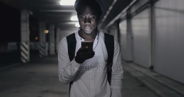 Hombre africano en sombrero usando teléfono inteligente pasear en el aparcamiento por la noche — Vídeo de stock