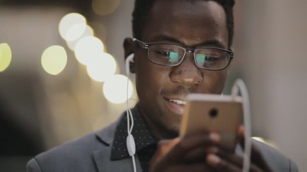 Joven estudiante africano en traje clásico escuchando música con auriculares al aire libre — Vídeos de Stock