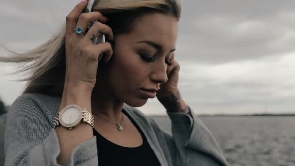 Femme écouter de la musique dans les écouteurs debout à la rivière, le vent souffle ses cheveux, automne journée nuageuse froide . — Video