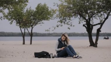 Kadın soğuk sonbahar gün kumsalda oturuyor ve smartphone kullanarak