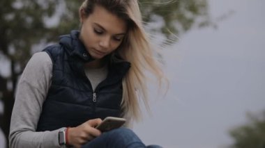 Kadın soğuk sonbahar gün kumsalda oturuyor ve smartphone kullanarak