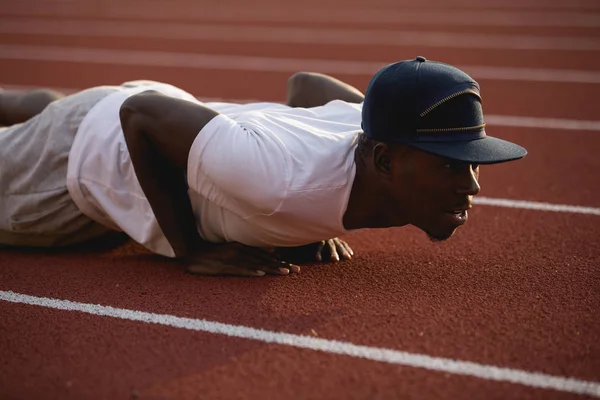 African male sportsman in a cap training pushups on stadium track — Stock Photo, Image