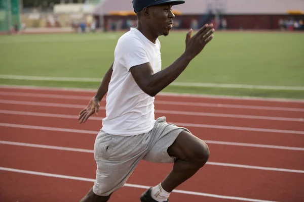 African male sportsman in a cap fast running short distance on stadium track, he trains speed