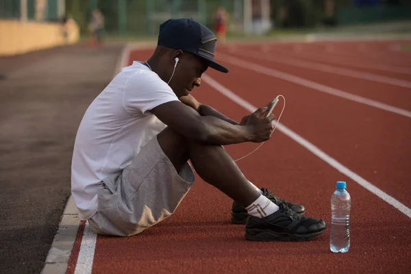 African american male sportsman sit on stadium track and listen music after hard workout — Stock Photo, Image