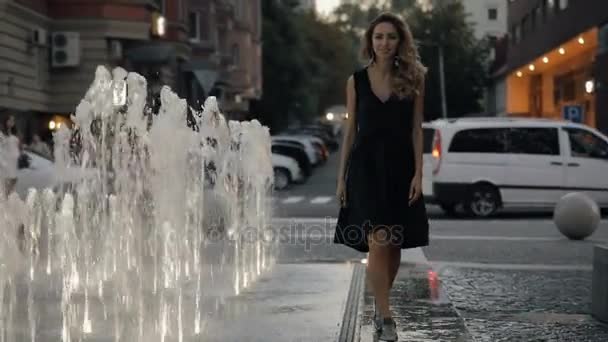 Jolie femme avec de beaux cheveux marchant dans une rue de ville du soir près de la fontaine, au ralenti — Video