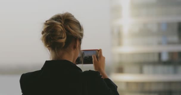 Mujer joven fotografiando paisaje urbano y construyendo en su teléfono inteligente al atardecer — Vídeo de stock