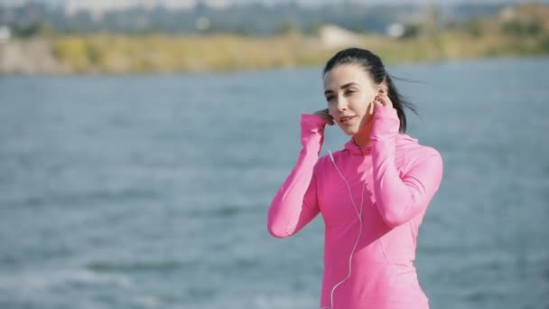 Hübsche Sportlerin in pinkfarbener Sportbekleidung beim Joggen im Freien in der Nähe von Fluss oder Meer, gesundes Lifestyle-Konzept. Zeitlupe. sie misst die Zeit auf der Stoppuhr. — Stockvideo
