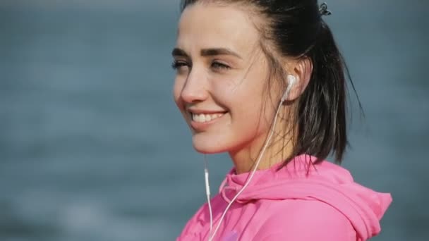 Retrato de cerca de la deportista bonita en ropa deportiva rosa sonriendo mirando a la cámara cerca del río o el mar, concepto de estilo de vida saludable. Movimiento lento . — Vídeo de stock