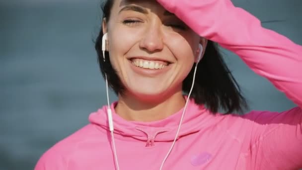 Retrato de cerca de la deportista bonita en ropa deportiva rosa sonriendo mirando a la cámara cerca del río o el mar, concepto de estilo de vida saludable. Movimiento lento . — Vídeo de stock