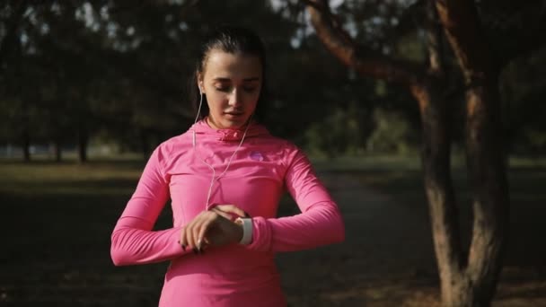 Όμορφη αθλήτρια σε ροζ αθλητικά ρούχα τζόκινγκ σε εξωτερικούς χώρους, έννοια του υγιεινού τρόπου ζωής. Αργή κίνηση. Βλέπει το χρόνο στο χρονόμετρο. — Αρχείο Βίντεο
