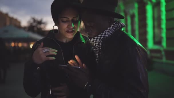 Lento movimiento de la mujer blanca y el hombre negro pareja mirando hacia abajo a la pantalla en el teléfono inteligente por la noche. Sonríen, ríen . — Vídeo de stock