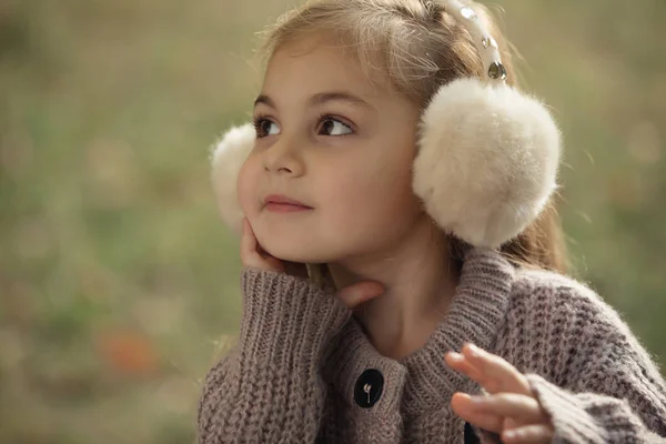 Портрет милой маленькой девочки в осенней одежде и наушниках, которые греют ей уши, сидящей на траве в городском парке — стоковое фото
